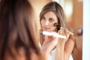 Nuori nainen käyttää aurinkopuuteria peilin edessä, keskittyneenä meikkaamiseen ja kauniiseen lopputulokseen.