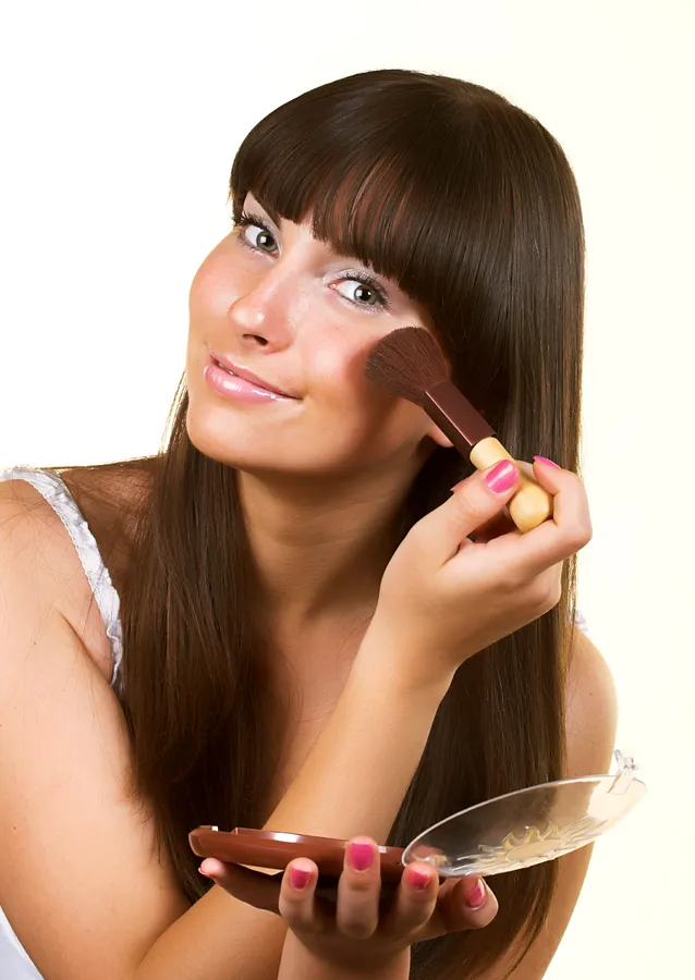 Nainen käyttää aurinkopuuteria poskilleen katsoen peiliin, hiukset pitkät ja mustat.