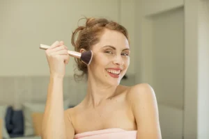 Nuori nainen käyttää meikkisivellintä aurinkopuuterin levittämiseen kasvoille, hymyillen iloisesti.