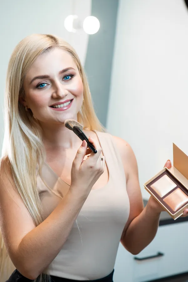 Nuori nainen hymyilee ja käyttää meikkisivellintä highlighter-paletista kauniin hehkun saavuttamiseksi.