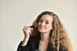 Nainen meikkaa pehmeällä siveltimellä highlighteria poskilla, taustalla vaalehtava seinä.