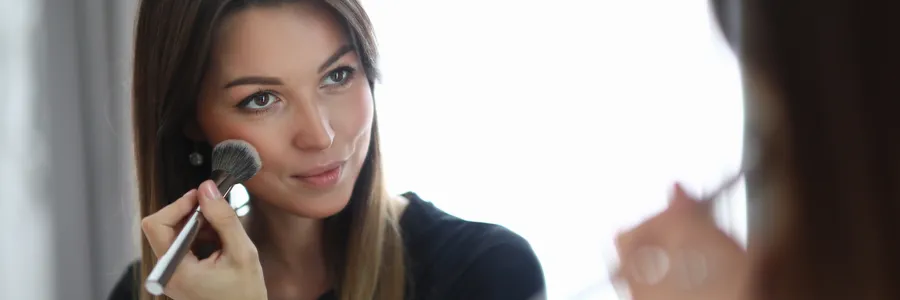 Nuori nainen siveltimellä levittää highlighteria poskilleen peilin edessä, luoden kauniin hehkun.