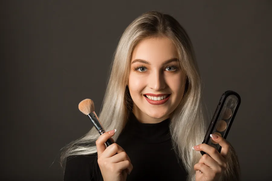 Hymyilevä nuori nainen pitää kädessään highlighteria ja meikkisivellintä, valmistautuen kauniiseen meikkiin.