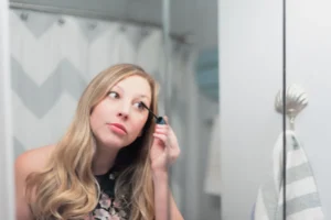 Nuori nainen käyttää ripsiväriä peilin edessä, keskittyneenä ilmeeseen. Hänellä on pitkät, vaaleat hiukset ja päällään kukkakuosinen toppi. Taustalla näkyy kiharapeite ja kylpyhuoneen sisustusta.