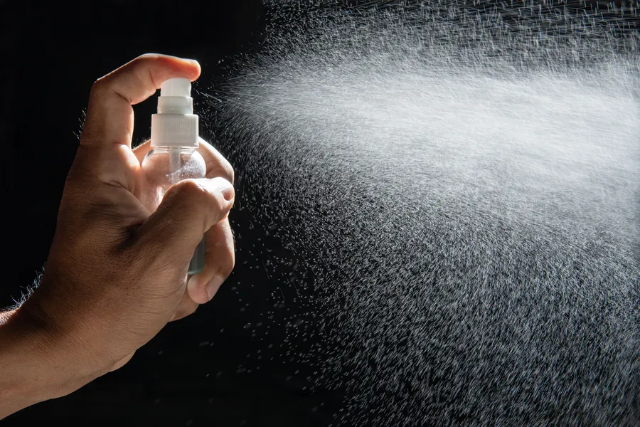 Käsi pitää suihkepulloa, josta suihkuaa setting spray'n hienoa sumua mustalla taustalla.