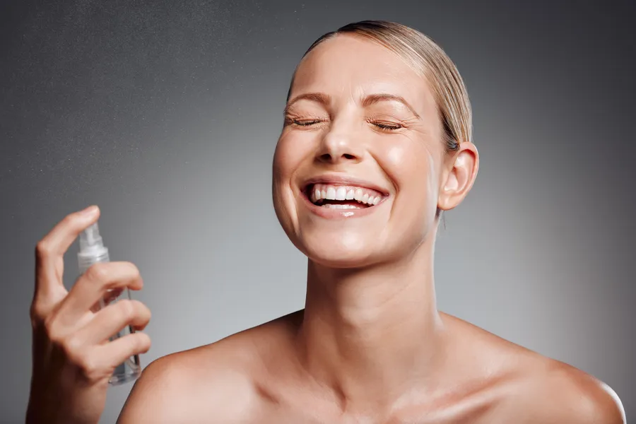 Nainen hymyilee levittäessään setting spray:ta kasvoilleen, tuoden luonnollista hehkua.