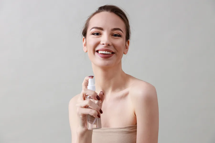 Iloinen nainen käyttää setting spray'ta, hymyilee ja näyttää kauniilta. Taustalla neutraali seinä korostaa hänen kauneuttaan.